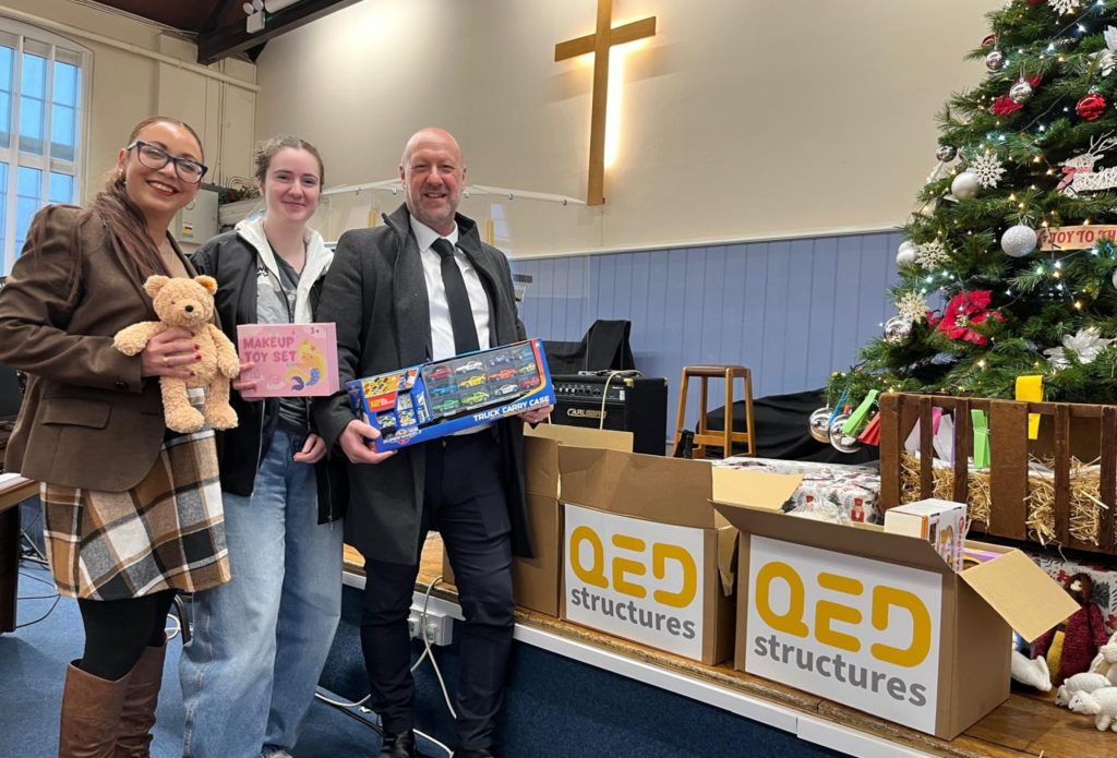 2 Woman and 1 Man stood together in front of a Christmas tree with boxes of gifts placed in front, inside a church.