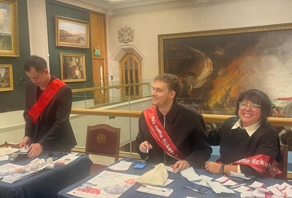 2 men and 1 woman sat at a table with smart black clothes and red sachets, in front of some historical looking paintings