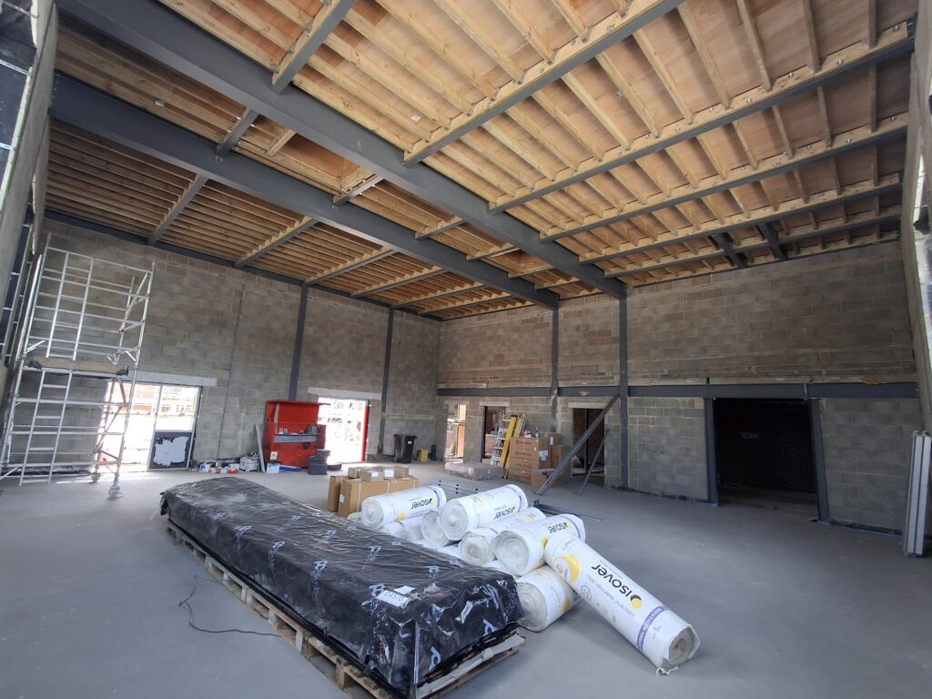 Large open interior under construction featuring exposed steel beams, timber floor decking, blockwork walls, and building materials stored on the concrete floor.